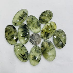 May include: A collection of ten green and black oval-shaped gemstone cabochons. The stones have a translucent appearance with black inclusions that resemble thin lines or threads.