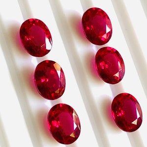May include: Six oval-shaped, deep red gemstones on a white background. The gemstones are arranged in two rows of three.