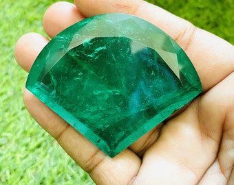 Puede incluir: Una gran gema verde esmeralda facetada en forma de semicírculo. La gema se sostiene en una mano, con un fondo verde de hierba.