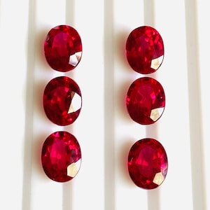 May include: Six oval-shaped, deep red gemstones on a white background. The gemstones are arranged in two rows of three.