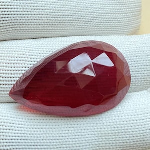 May include: A deep red pear-shaped gemstone with a faceted cut. The gemstone is on a white fabric background.