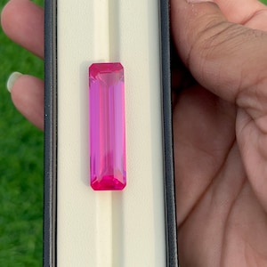 Pode incluir: Uma gema rosa vibrante de forma retangular, colocada em uma caixa de exibição branca com borda preta.