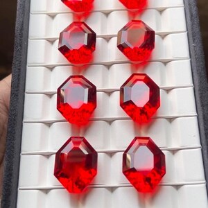 May include: Eight faceted, deep red gemstones arranged in a display case. Each gem has a multi-sided shape, reflecting light. The gems are set against a white background within a black case, creating a striking contrast.