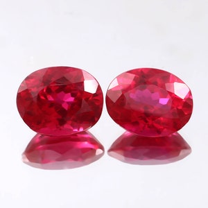 May include: Two oval-shaped, red gemstones with a high polish. The gemstones are set against a white background.