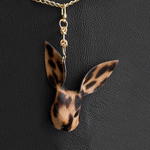 May include: A pendant necklace with a rabbit head design, displaying a brown and black spotted pattern. The pendant hangs from a gold-coloured chain. The background is black. A distinctive piece of jewellery.