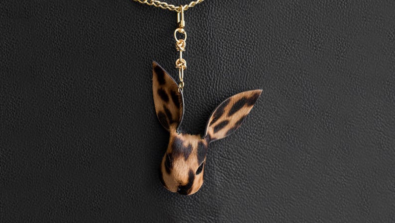 May include: A pendant necklace with a rabbit head design, displaying a brown and black spotted pattern. The pendant hangs from a gold-coloured chain. The background is black. A distinctive piece of jewellery.
