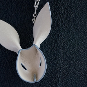 May include: A cream leather bunny mask with long ears and eye holes, hanging from a silver chain. The mask has a light blue trim and is set against a dark blue textured background.