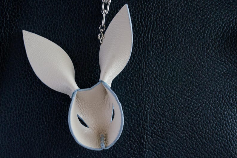 May include: A cream leather bunny mask with long ears and eye holes, hanging from a silver chain. The mask has a light blue trim and is set against a dark blue textured background.