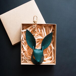 May include: A teal leather rabbit head keychain with a gold-colored clasp, presented in a kraft paper gift box filled with shredded paper. The box is open, revealing the keychain against a dark background.