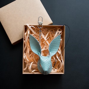 May include: A pale blue leather bunny keyring with a silver-tone clasp, displayed in a brown gift box with shredded paper. The bunny features detailed ears and a textured surface, set against a black background.