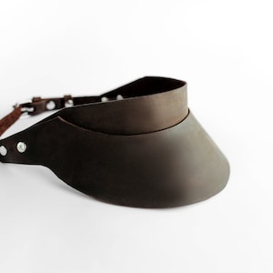 May include: A brown leather visor with a strap and buckle fastening. The visor is made of two layers of leather, with the top layer being slightly smaller than the bottom layer.