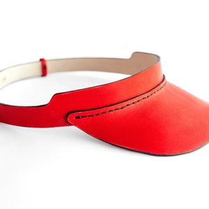 May include: A red leather visor with a stitched seam. The visor is curved and has a wide brim.