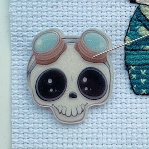 May include: A white skull with large black eyes and brown goggles. The skull is decorated with glitter and has a small smile. The skull is on a white background.