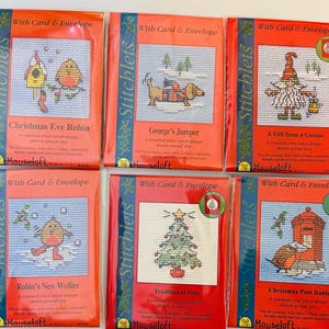 May include: Six cross-stitch kits with red packaging, each featuring a different Christmas-themed design. The kits include designs like a gnome, a Christmas tree, and a robin. Each kit is labelled "Stitchlets" and includes a card and envelope.
