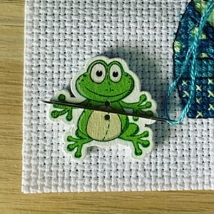 May include: A green frog-shaped wooden button with a black buttonhole, threaded with a needle and white thread, on a white Aida cloth.