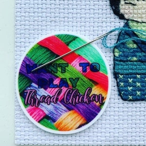 May include: A round sticker with a colourful yarn background and the text "Want to play Thread Chicken" in a white circle. A needle is threaded with blue yarn and is partially inserted into the sticker.