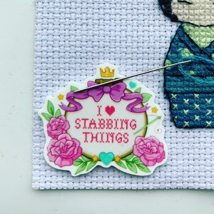 May include: A white cross-stitch embroidery hoop with a pink and purple floral design. The hoop has a pink heart and the text "I ♥ STABBING THINGS".