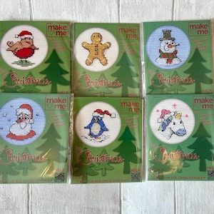 May include: Six individual Make Me cross-stitch kits for Christmas, each in a green package with a circular window. Designs include a robin, gingerbread man, snowman, Santa Claus, penguin, and unicorn, all featuring festive holiday themes.