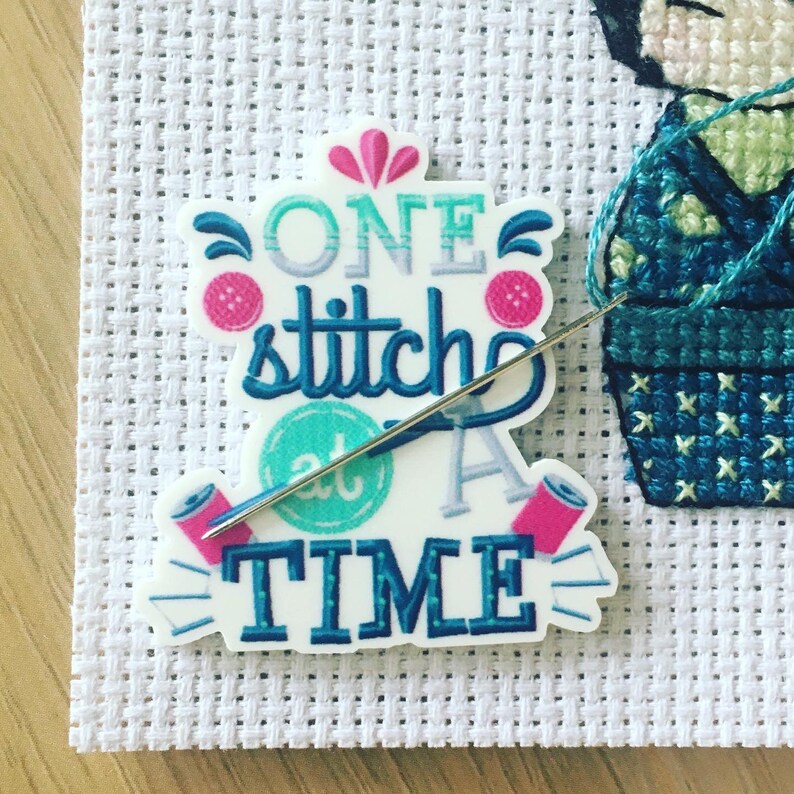 Może przedstawiać: Biała naklejka z tekstem "ONE stitch at a TIME" w niebieskich i zielonych literach. Naklejka ma wz&oacute;r r&oacute;żowego kwiatu i igłę z nitką przechodzącą przez nią.