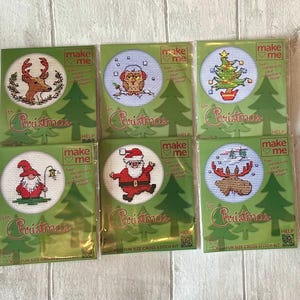 May include: Six individual Make Me cross stitch kits for Christmas, each featuring a circular design including a reindeer, owl, Christmas tree, gnome, Santa Claus, and moose. The green packaging displays the text Make Me and for Christmas.