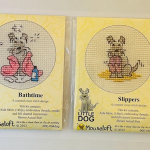 May include: Two cross-stitch kits with dog designs. One shows a dog in a bathtub with pink towels and a blue bottle, labelled "Bathtime." The other shows a dog wearing pink slippers, labelled "Slippers."