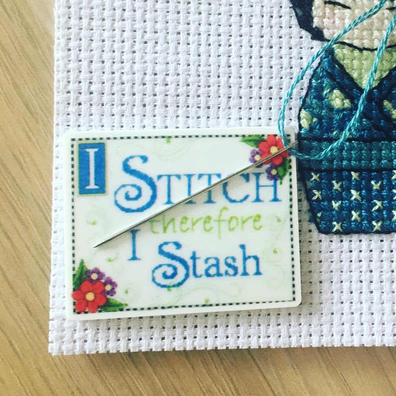 Może przedstawiać: Biały projekt haftu krzyżykowego z igłą i nicią. Projekt zawiera małą, kolorową naklejkę z tekstem "I Stitch therefore I Stash".
