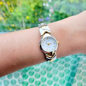 May include: A silver and gold two-tone wristwatch with a white face and Roman numeral hour markers. The watch face has the text "Anne Klein" and "Swiss" printed on it.