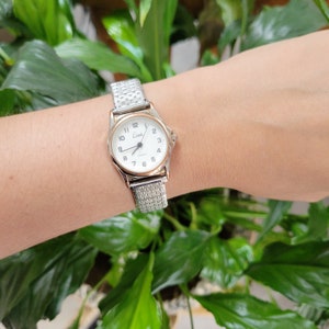 May include: A silver-toned Limit quartz watch with a white face and black numerals is displayed. The watch has a mesh band and is worn on a wrist. The watch face has a gold-colored bezel. The background is a lush green plant.
