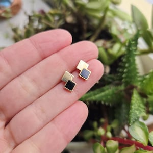 May include: A pair of gold-colored square stud earrings, each with a black square inlay. The earrings are displayed on a hand, with a blurred background of green plants.
