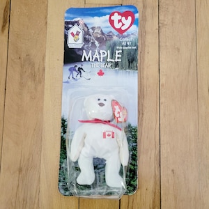 May include: White Ty Beanie Baby plush toy named "Maple the Bear" with a red ribbon and a Canadian flag patch. The toy is packaged in a plastic bag with a Ty tag and a cardboard backing with the words "Collect All 4!" and "While supplies last."