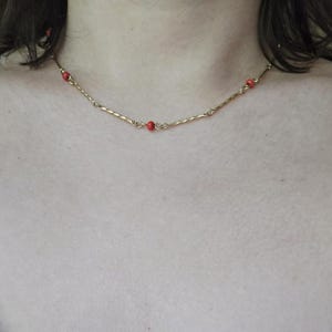 May include: A delicate gold necklace featuring small red beads. The necklace has a textured, twisted design and is worn around the neck. The necklace is a simple, elegant piece of jewelry.