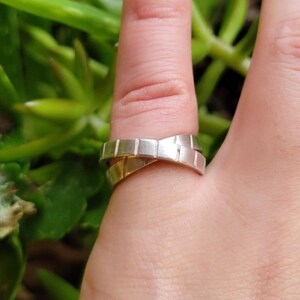 Può includere: Un anello d'argento con un design a croce. L'anello è composto da due bande intrecciate e ha una superficie testurizzata.