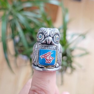 May include: A silver-toned owl-shaped ring with blue gemstone eyes. The ring features a blue and red emblem with the word "MALLORCA". The ring is worn on a finger.
