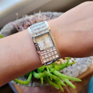 May include: Silver-toned quartz watch with a rectangular face and a band adorned with square crystals. The watch face is cream-colored with black numerals and hands. The watch is on a wrist.