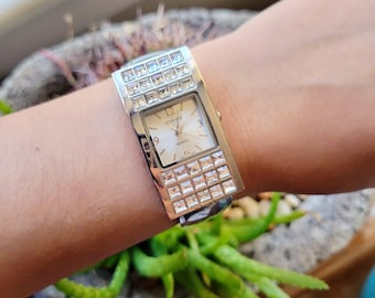 May include: Silver-toned quartz watch with a rectangular face and a band adorned with square crystals. The watch face is cream-colored with black numerals and hands. The watch is on a wrist.