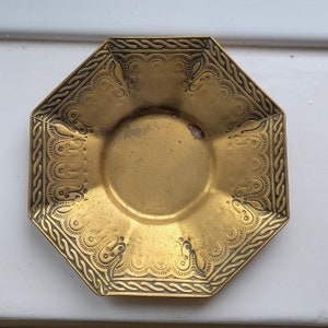 Può includere: Un piatto ottagonale in metallo dorato con un design intricato attorno al bordo. Il piatto ha un centro rialzato e una superficie liscia leggermente usurata.