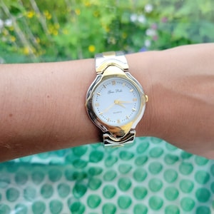 May include: A silver and gold two-tone watch with a white face and black hands. The watch face has Roman numeral hour markers and the text "Gino Palli" and "QUARTZ".