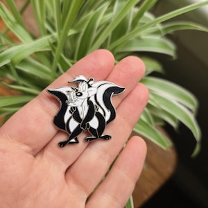 May include: A black, white, and silver enamel pin featuring the cartoon character Pepe Le Pew. The pin depicts the skunk with his arms crossed, with a black and white striped design. The pin is held in a hand.