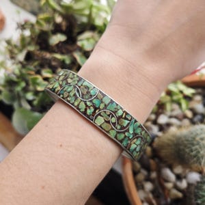 May include: A silver-toned bracelet with a mosaic design of turquoise-colored stones and dark brown accents. The bracelet features a repeating pattern of silver-toned circles. The jewelry is displayed on a wrist.