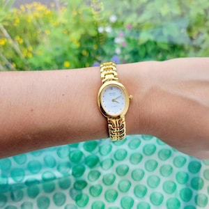 Op de afbeelding: Een goudkleurig horloge met een witte wijzerplaat en een goudkleurige band. De wijzerplaat van het horloge heeft zwarte Romeinse cijfers als uurmarkeringen en zwarte wijzers. Op de wijzerplaat van het horloge staat ook "SEIKO QUARTZ" gedrukt.