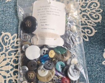 Op de afbeelding: Een doorzichtige plastic zak gevuld met een verscheidenheid aan knopen in verschillende maten, kleuren en materialen. De knopen omvatten zwarte, witte, grijze en iriserende opties. Een label luidt "Fullfashioned Sweater made of 100% Acrylic Fiber...HONG KONG."