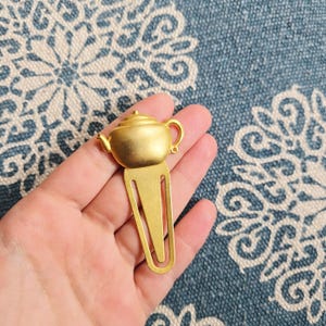 Può includere: Un segnalibro in metallo color oro a forma di teiera. La teiera ha un piccolo manico e beccuccio e si trova sopra una clip per segnalibro rettangolare. Il segnalibro è tenuto in mano su uno sfondo blu e bianco.