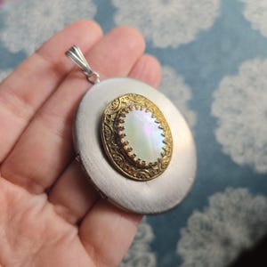 May include: A silver-toned locket with a decorative gold-colored border and a pearlescent center. The locket has a hinged design and a silver bail for a chain. The locket is round and has a vintage aesthetic.