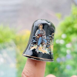 May include: A black and gold ceramic thimble with a painted image of a man in traditional clothing playing a musical instrument. The thimble has a white rim and is decorated with a blue and white pattern.