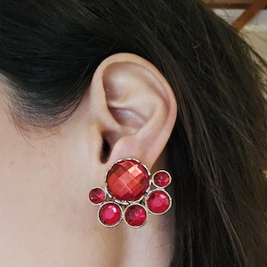 May include: A close-up of a red gemstone earring. The earring features a large, faceted, round red stone surrounded by smaller red stones. The setting is gold-colored, and the earring is clipped to an ear.