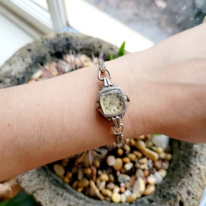 Könnte beinhalten: Eine silberne Armbanduhr mit quadratischem Zifferblatt und einem zarten Kettenarmband. Das Zifferblatt hat schwarze römische Ziffern und einen kleinen Sekundenzeiger.