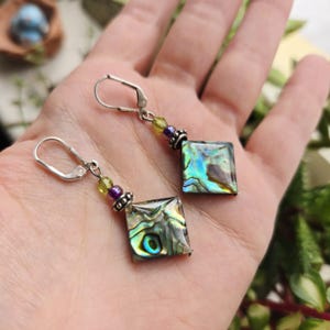 May include: A pair of square abalone shell earrings with silver-tone lever-back closures. The earrings feature iridescent squares with green, purple, and yellow beads. The earrings are held in a hand.