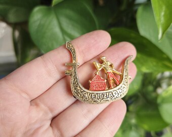 May include: A gold-toned metal brooch in the shape of a Venetian gondola. The gondola features a red and gold figure of a gondolier and a red structure. The word "VENEZIA" is inscribed on the boat.