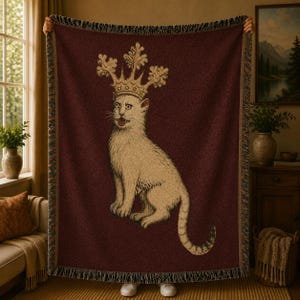 May include: A woven throw blanket featuring a deep burgundy background with a central illustration of a light-colored cat wearing a large, ornate gold crown. The cat is depicted in a seated position with its mouth slightly open, and the blanket has a fringed edge.