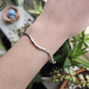 May include: A silver-coloured, wavy cuff bracelet with a hammered texture, worn on a wrist. The background features green plants and a small decorative item.